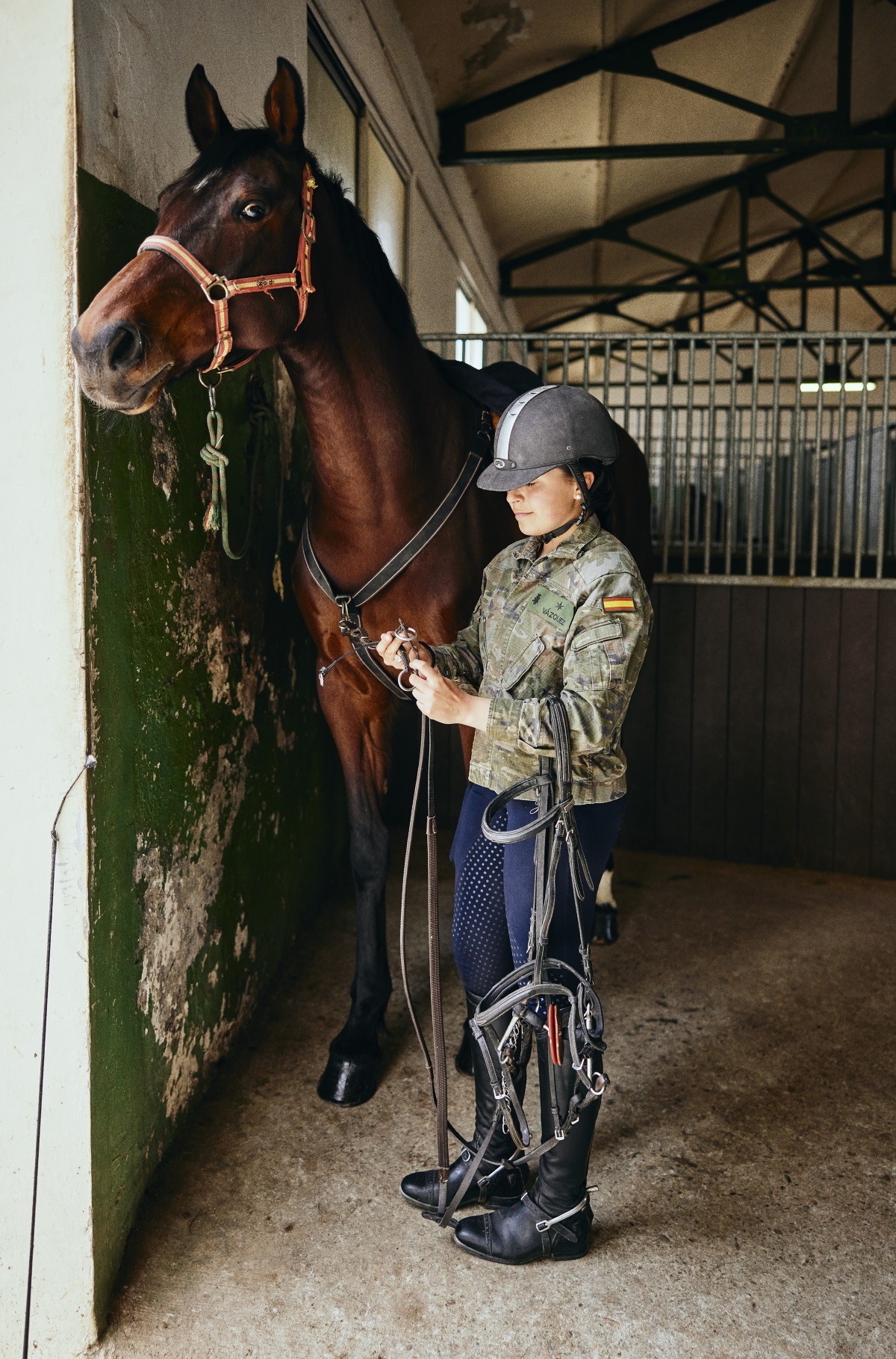 La dama cadete de 3er curso, Elena Martínez Vázquez, en las cuadras poniendo las bridas a su caballo de saltos. Lleva la chaqueta de instrucción o "mimeta" y el pelo recogido en una trenza.