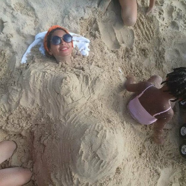 Beyonce tumbada en la playa con arena encima haciendo forma de mujer embarazasda.